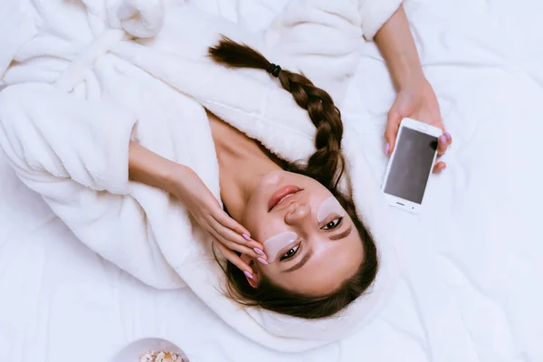 Mulher deitada na cama aplicando creme no rosto — Fotografia de Stock