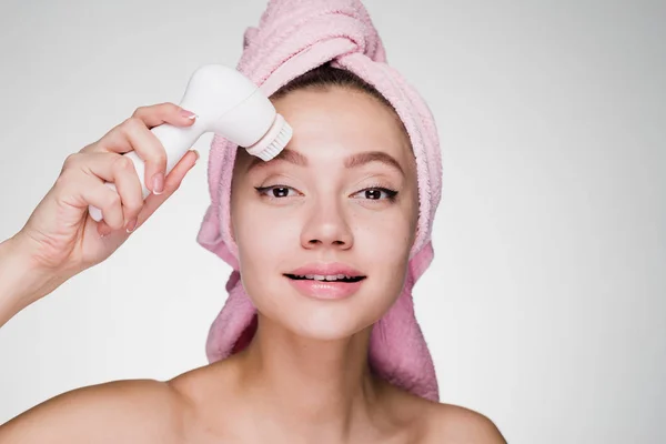 Vrouw met een handdoek op haar hoofd reinigt het gezicht met een borstel voor diepe reiniging — Stockfoto