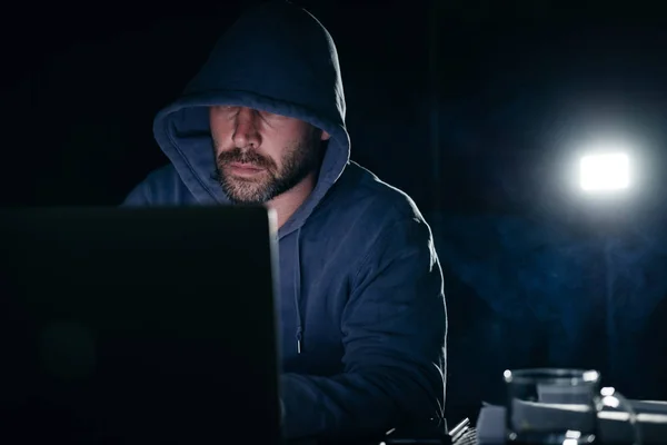 Pirate masculin dans un sweat-shirt avec une capuche assise à une table et regardant un ordinateur portable dans une pièce sombre — Photo