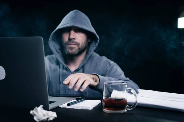 Αρσενικό χάκερ σε ένα φούτερ με κουκούλα κάθεται σε ένα τραπέζι και πίνοντας τσάι — Φωτογραφία Αρχείου