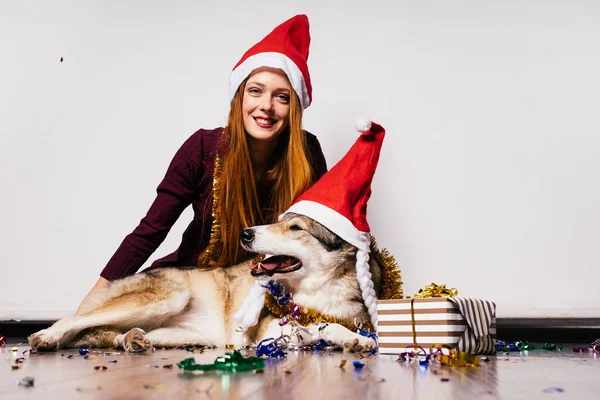 Happy woman in a New Year's cap sits next to a dog on the background of gifts — Stock Photo, Image