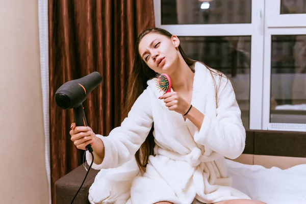 Vrouw zittend op het bed en waait haar haren met een haardroger — Stockfoto