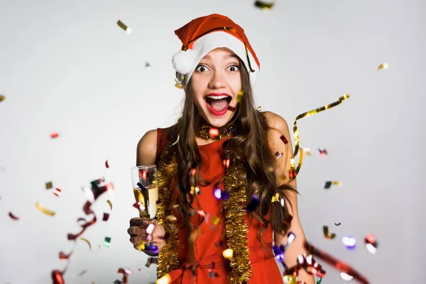 Happy νεαρό κορίτσι γιορτάζει το νέο έτος, στο κεφάλι της ένα κόκκινο σκουφάκι σαν τον Άγιο Βασίλη, πολλά πολύχρωμα κομφετί — Φωτογραφία Αρχείου