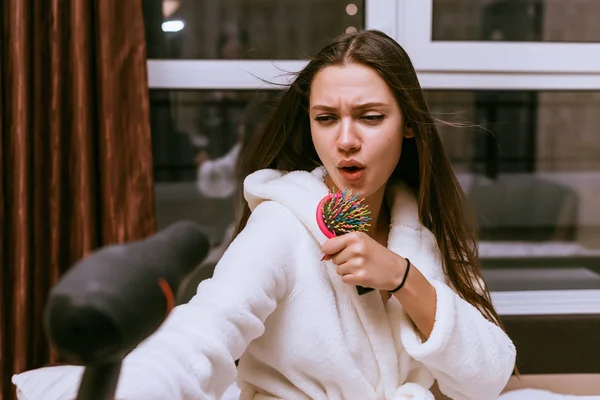 Divertida joven en una bata blanca seca su cabello con un secador de pelo y canta en un peine — Foto de Stock