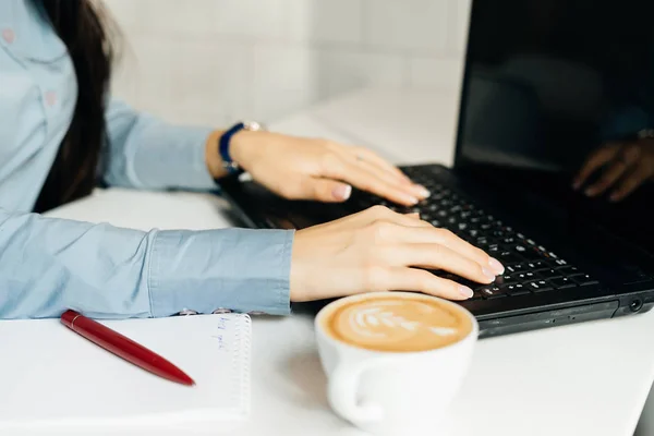 Freelancer menina em uma camisa azul funciona em um laptop, na mesa fica café — Fotografia de Stock