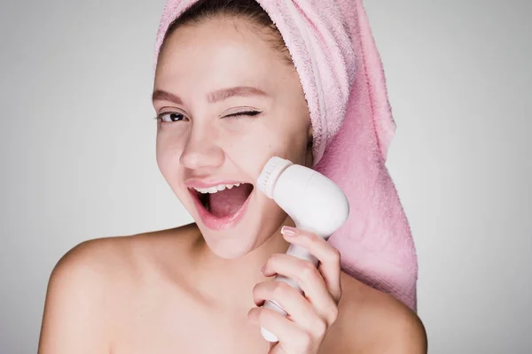 Μια έκπληξη γυναίκα με μια πετσέτα στο κεφάλι της καθαρίζει το δέρμα με ένα βουρτσάκι για βαθύ καθαρισμό του προσώπου — Φωτογραφία Αρχείου