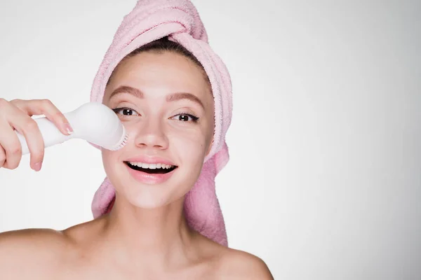 Femeie fericită cu un prosop pe cap curăță pielea cu o perie pentru o față profundă de curățare — Fotografie, imagine de stoc