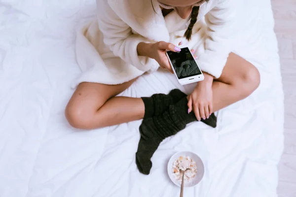 Женщина сидит на кровати и держит телефон в руках. — стоковое фото