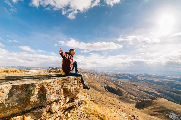 Mulher feliz turista senta-se em um penhasco no fundo das montanhas em um dia ensolarado — Fotografia de Stock