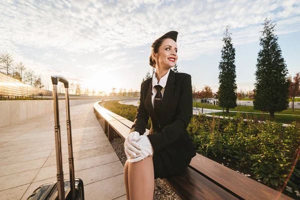 Letuška šťastná žena v uniformě sedí vedle kufr na pozadí oblohy — Stock fotografie