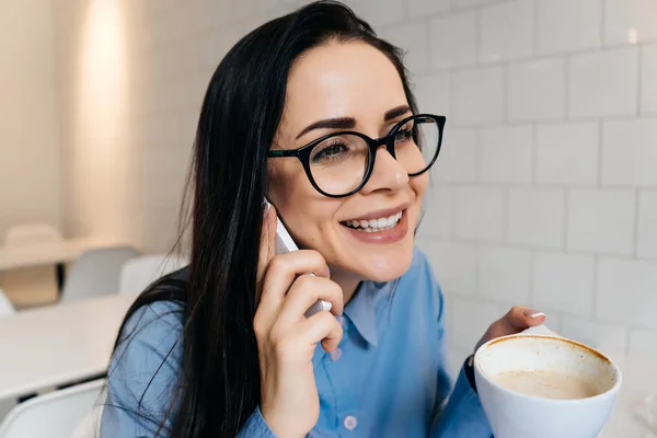 happy woman sitting at table in office drinking coffee and talking on the phone