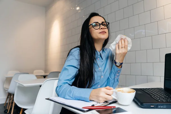 woman sitting at table in office drinking coffee and eating sandwich