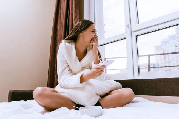 Happy woman sits on the bed and looks out the window — Stock Photo, Image