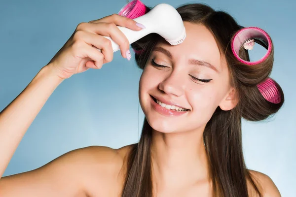 Donna felice con bigodini sulla testa pulisce la pelle con un pennello per una pulizia profonda del viso — Foto Stock