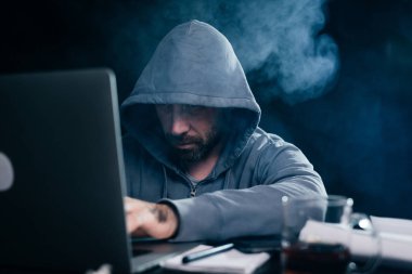 bir adam hacker bir kukuleta ile bir kazak'ı bir masada oturuyor ve bir şey bir dizüstü bilgisayarda yazarak