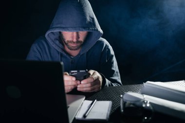 bir kazak bir kukuleta ile erkek hacker masada oturur ve telefona benziyor