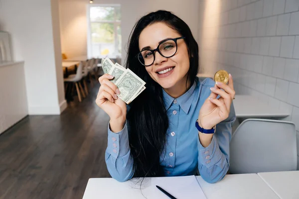 Mujer feliz sentada en una oficina en una mesa y sosteniendo una moneda criptográfica —  Fotos de Stock