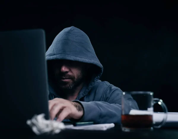 Mannelijke hacker in een sweatshirt met capuchon zitten aan een tafel en kijken naar de laptop — Stockfoto