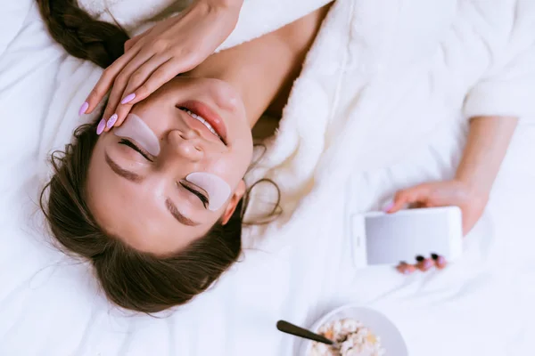 Mujer feliz poner parches bajo los ojos y se encuentra en la cama —  Fotos de Stock