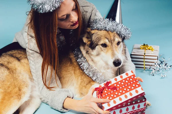 Woman in a festive hat hugs a big dog and opens a gift — Stock Photo, Image