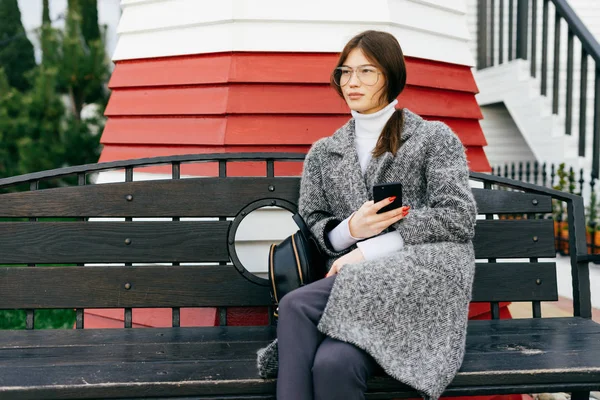 Une jeune fille forte dans un manteau gris à la mode et des lunettes s'assoit sur un banc dans la rue, tient un smartphone — Photo