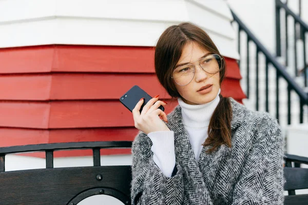 Bella ragazza elegante in occhiali alla moda e un cappotto grigio è seduto sulla strada, tenendo uno smartphone in mano — Foto Stock