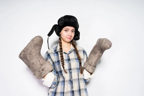 Lustige junge Russin mit schwarzem Hut und Ohrenklappen hält graue Filzstiefel in den Händen — Stockfoto