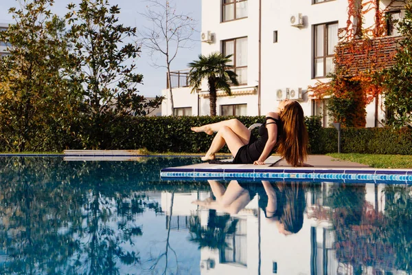 Una atractiva mujer rica en un vestido negro disfruta relajarse junto a la piscina, la vida de lujo y el pelo largo — Foto de Stock
