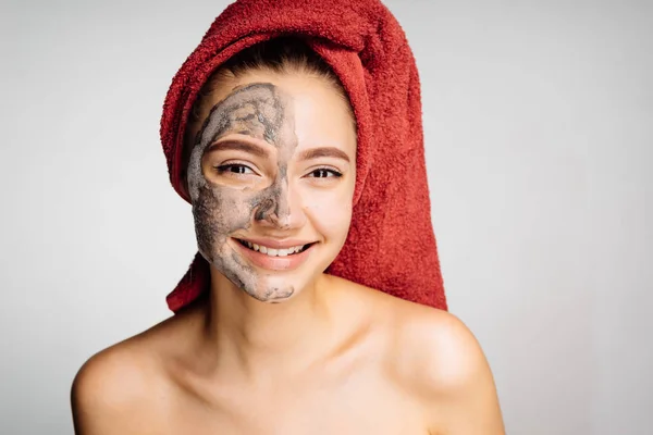 Une jolie jeune fille avec une serviette sur la tête s'occupe de son visage, met un masque d'argile sur la moitié de son visage, sourit — Photo
