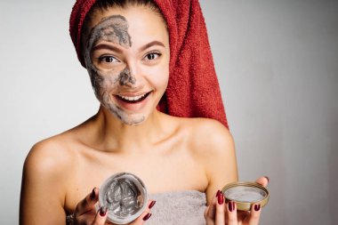 mutlu genç kız başında kırmızı bir havlu ile yararlı kil maskesi yüzünde uygular.