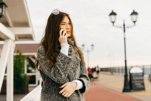 Cute long-haired girl in gray coat talking on the phone, walking along the street — Stock Photo, Image