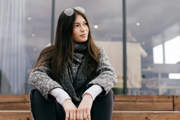 Bella giovane ragazza elegante in un cappotto grigio e occhiali è seduto sulla strada sui gradini, pensando a qualcosa — Foto Stock