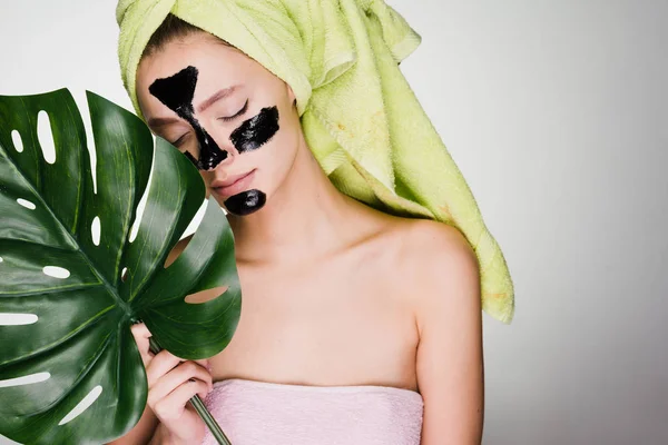 Joven con una toalla verde en la cabeza disfrutando de un spa, bajo los ojos de manchas negras, sostiene una hoja verde — Foto de Stock