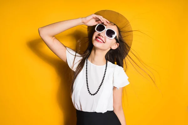 Menina elegante feliz em um chapéu na moda e óculos de sol posando contra um fundo de parede amarelo — Fotografia de Stock