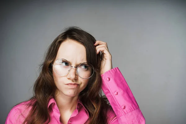 Joven chica oficina trabajador en un rosa camisa y gafas pensativamente rasca su cabeza — Foto de Stock