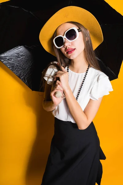 Menina elegante feliz em chapéu na moda e óculos de sol posando no fundo preto e amarelo — Fotografia de Stock
