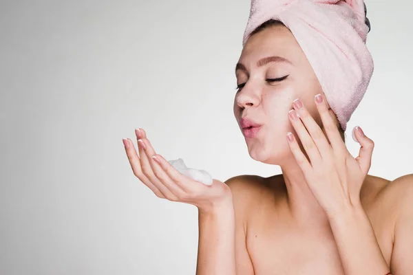 Uma menina bonito com uma toalha rosa na cabeça aplica uma espuma de limpeza em seu rosto — Fotografia de Stock