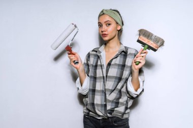 ekose gömlekli bir genç kız evinde onarım yapıyor, bir fırça ve bir silindir duvarları boyama için tutuyor