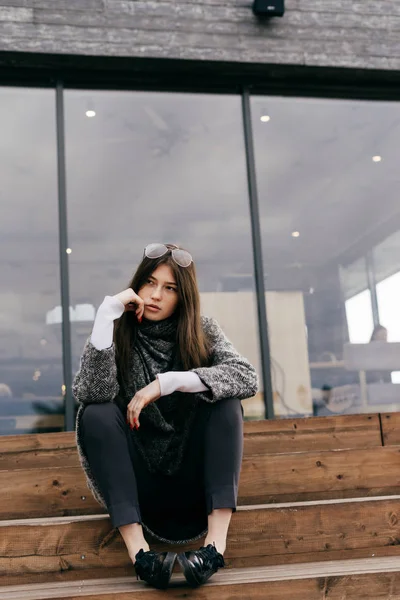 Стильна молода темноволоса дівчина в сірому светрі і окулярах сидить на сходах на вулиці — стокове фото