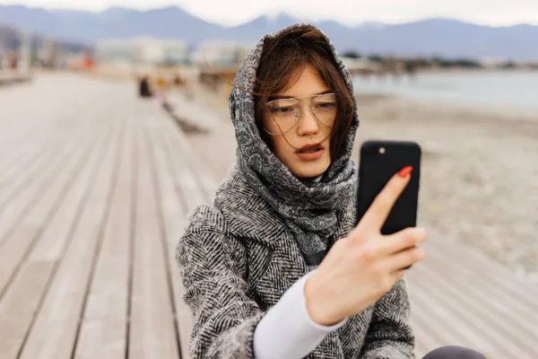 Stylish young girl with glasses walks and makes selfie on her smartphone — Stock Photo, Image