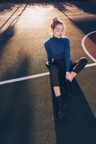 Elegante ragazza bionda in un maglione blu si siede su un campo sportivo al sole, gli occhi sono chiusi — Foto Stock