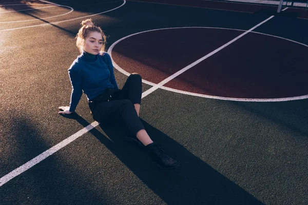 Стильна приваблива дівчина в синьому светрі сидить на спортивному полі позує — стокове фото