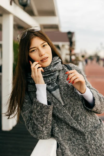 Elegante bella ragazza dai capelli scuri in cappotto grigio parlando al telefono, passeggiando per la città — Foto Stock