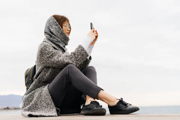 Stylish young girl with glasses and a gray coat sits on the ground and takes pictures of beautiful nature — Stock Photo, Image