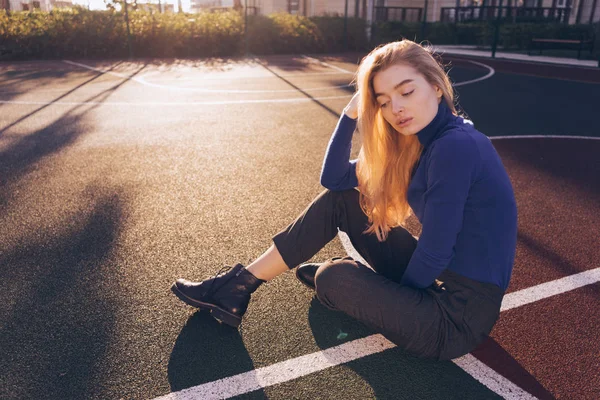 Bella giovane bionda dai capelli lunghi ragazza in maglione blu si siede sul campo sportivo in città, posa, gli occhi chiusi — Foto Stock