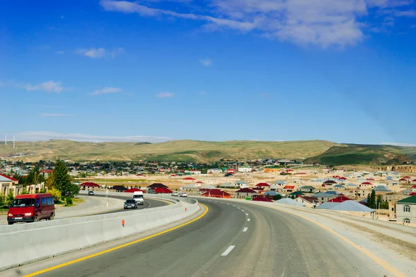 On the highway cars are passing by a small city against the backdrop of high mountains and a blue sky — Stock Photo, Image