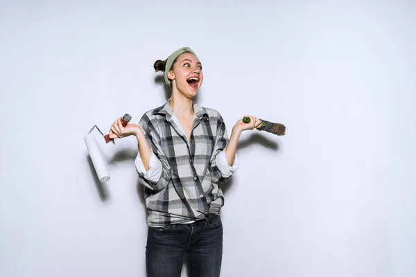 Счастливая смешная девушка в рубашке делает ремонт в своей квартире, держа ролик и кисть для покраски стен — стоковое фото