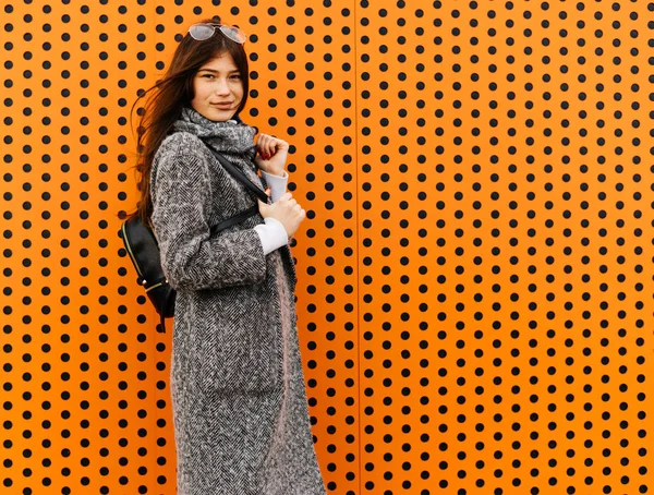 Élégante fille mignonne dans un manteau gris à la mode posant contre un mur orange inhabituel, style de rue — Photo