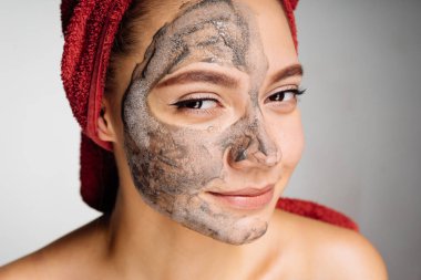 başında kırmızı bir havlu ile sevimli bir genç kız bir kil maskesi yüzü, yarısı için uygulanan gülümseyen