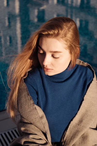 Carina ragazza bionda in maglione blu siede vicino alla piscina, al sole, gli occhi sono chiusi — Foto Stock